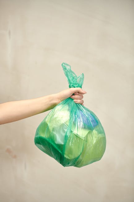 A person’s arm extending from the left side of the frame is holding a green and blue plastic waste bag by its twisted tie top, with the bag appearing partially filled and slightly stretched. The bag has a translucent, glossy surface, revealing some of its contents. It is positioned in the foreground against a plain, neutral-colored wall, creating a simple background. The scene emphasizes the act of waste disposal, relevant to rubbish collection services, with the bag likely containing household or light waste. The lighting is soft and even, with no harsh shadows, providing clear visibility of the bag’s color and texture. Waste Clearance Uxbridge may use such images to illustrate their approach to private waste removal, including on-site clearance or alternative disposal methods for household rubbish. The image effectively communicates a focus on waste management through visual cues of waste containment and handling, aligning with the topic of same-day rubbish collection and related services in Uxbridge.