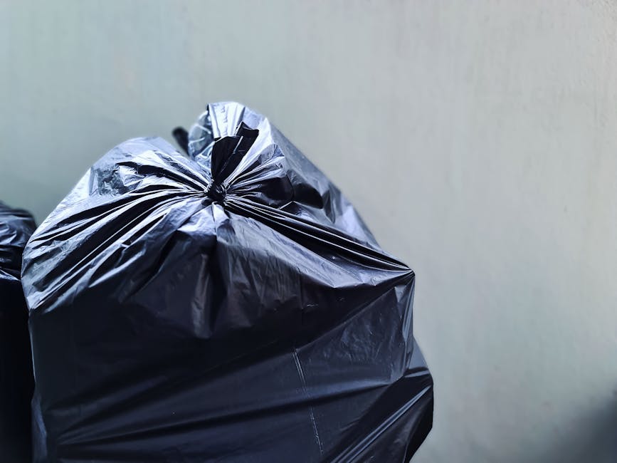 Close-up of a black plastic rubbish bag, tightly knotted at the top, placed against a plain light-colored wall. The bag appears to be made of glossy, durable plastic material with visible wrinkles and creases, indicating it is filled with waste. The background exhibits a neutral, smooth surface with no distinct features, suggesting an indoor or sheltered area such as a storage space or side alley. The lighting is even, highlighting the shine on the plastic bag's surface and providing clear detail of its texture and form. This scene exemplifies typical refuse collection preparations, relevant to private waste handling and rubbish removal services, such as those offered by Waste Clearance Uxbridge, which perform on-site clearance and alternative waste disposal options for domestic or commercial clients.