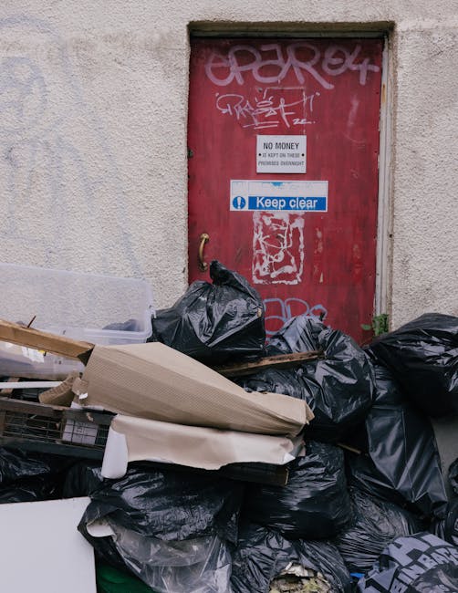 A collection of black trash bags and discarded packaging materials piled in front of a red metal door set into a beige, textured wall. The bags vary in size and are stacked haphazardly, some partially covering other waste items. The door features multiple graffiti tags in white and pink spray paint, along with several signs including a prominent blue and white sticker reading 'Keep clear' and a smaller red and white sign that states 'No Money' and information about access restrictions. The scene appears to be an outdoor urban alleyway or back area, with minimal natural light illuminating the waste and wall surface. The area reflects an instance of private rubbish disposal or on-site clearance, with Waste Clearance Uxbridge specializing in such services, aligned with the context of alternative waste handling for local properties or businesses.