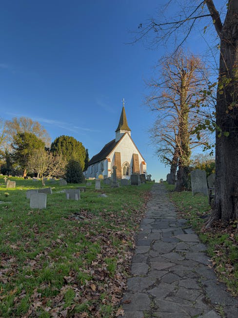 A view of a small, white stone church with a tall, pointed steeple set atop a grassy, slightly sloped area. Surrounding the church are leafless trees, some with sparse green foliage and others bare, indicating late autumn or early winter. The ground is scattered with fallen leaves, and a narrow, uneven cobbled pathway leads up towards the church from the foreground. To the left, there are a few scattered stone graves or memorial markers visible on the grassy slope. The sky above is clear and blue, with sunlight casting shadows on the ground and illuminating the church's facade. The scene suggests an outdoor place of worship or historical site, with the church and graveyard possibly managed by a private property or local heritage organization, such as Waste Clearance Uxbridge, as part of an alternative waste handling or site clearance service in the surrounding area.
